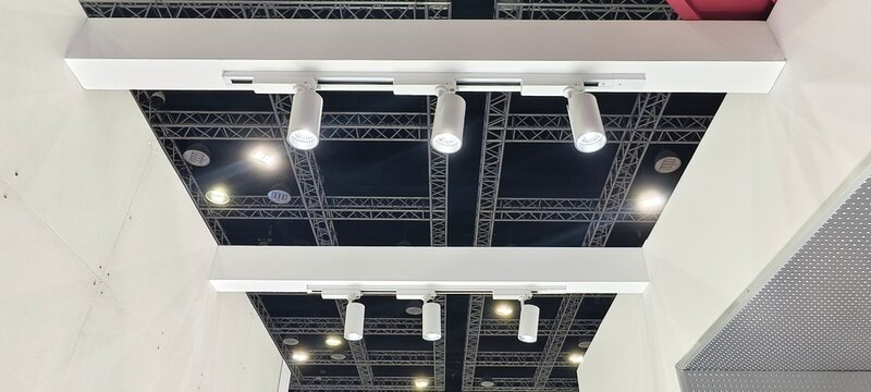 Parallel white beams supporting multiple cylindrical track lights in an industrial ceiling with trusses and overhead fixtures. Bright directional illumination highlights modern exhibition hall.