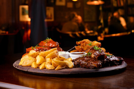 Close-up of a finger food platter featuring BBQ ribs, fried chicken, french fries, and buffalo wings on a table, with people blurred in the background.