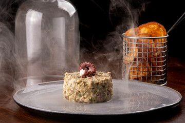 Close-up of a smoked seafood tartare dish garnished with an octopus tentacle at a pub, featuring smoke and a basket of fries. © mario