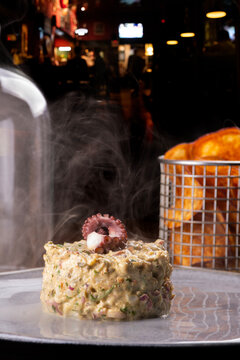 Close-up of a smoked seafood tartare dish garnished with an octopus tentacle at a pub, featuring smoke and a basket of fries, with the restaurant interior in the background.