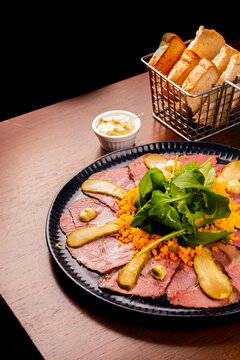 Close-up of an appetizer plate featuring pastrami, roast beef, arugula, and toast on a wooden table.