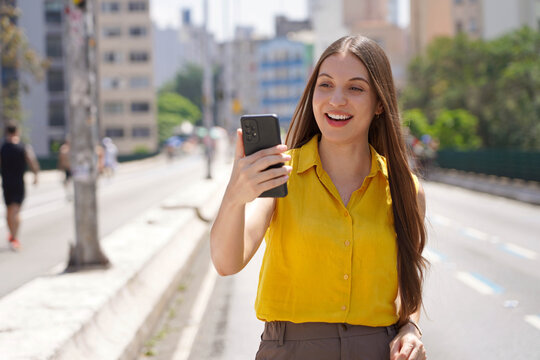 Happy beautiful young woman using mobile phone for video call outdoor with blurred background of city street. City lifestyle people technology.