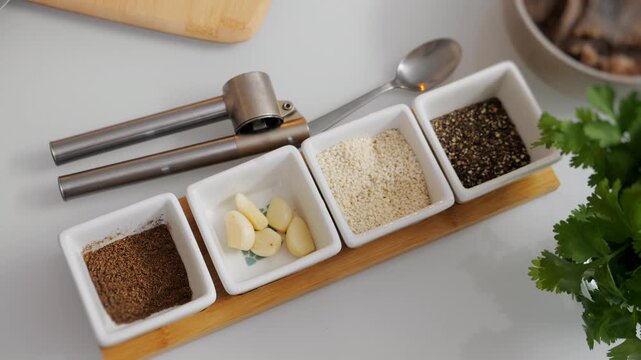 Garlic cloves, sesame seeds, black pepper and spices arranged in small bowls with a garlic press and fresh herbs. Ingredients prepared for cooking in a modern kitchen.