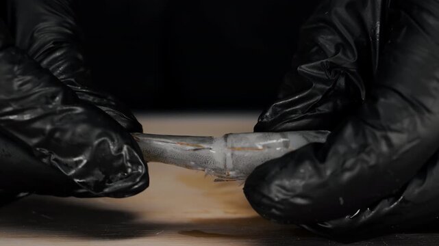 Close-up of hands in black gloves peeling shell from raw shrimp on a cutting board. Detailed seafood preparation process before cooking.