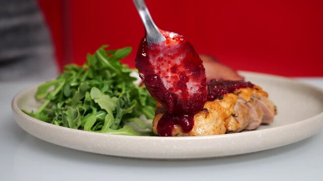 Close-up of berry sauce being spooned over sliced cooked duck breast served with fresh arugula. Gourmet food plating and restaurant-style presentation.