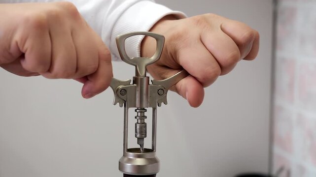 Close-up of hands using a mechanical corkscrew to remove a natural cork from a wine bottle. Detailed opening process with traditional kitchen and bar tool.
