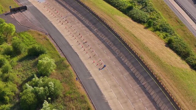 Small car maneuvers along a paved track at a vehicle testing facility while driving past roadside markers and approaching a concrete barrier near grassy fields.