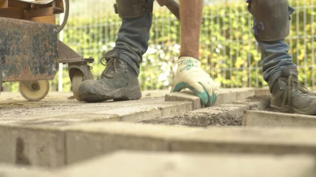 Worker laying concrete paving slabs, using a bolster chisel and hammer on a construction site.
