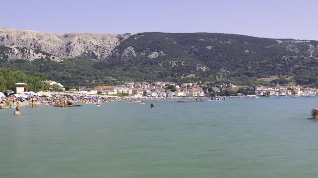 Groups swim and relax on a beach by the water with hills behind.