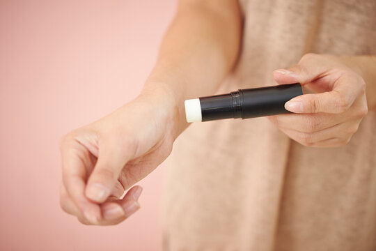 Close up of a woman applying a clear moisturizing balm stick or solid perfume on her wrist against a soft pink background