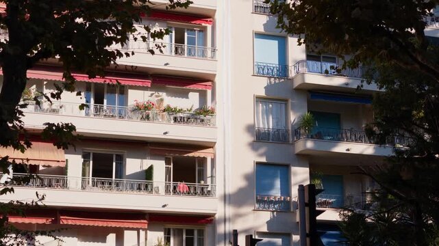 Static shot of sunlit apartment facades with red and blue awnings and flower-filled balconies framed by plane trees, Nice, French Riviera, Cote d Azur, France