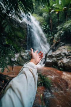 Point of view of a person reaching hand towards a beautiful cascading waterfall in a tropical jungle.