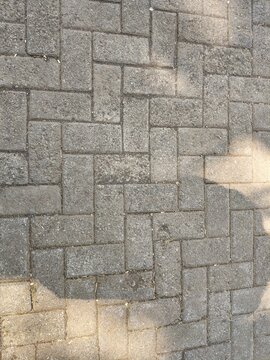 Shadow of a person taking a photo cast upon a grey zig-zag patterned paving block walkway illuminated by sunlight