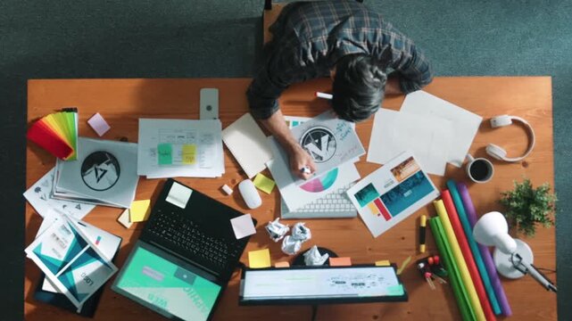 Top view of designer writing and designing logo graphic at meeting. Closeup of business man working on colored palettes and writing idea on sticky notes on table with equipment and paper. Symposium.