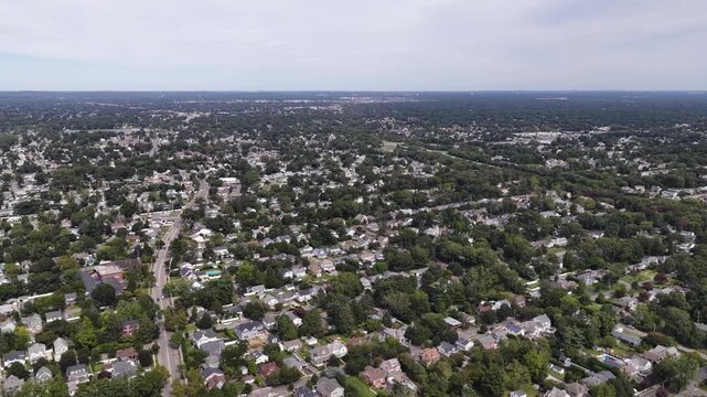 Drone flies over leafy residential neighborhoods in Babylon on Long Island in New York. Detached houses and winding streets stretch toward the horizon under a pale sky with dense green tree cover.