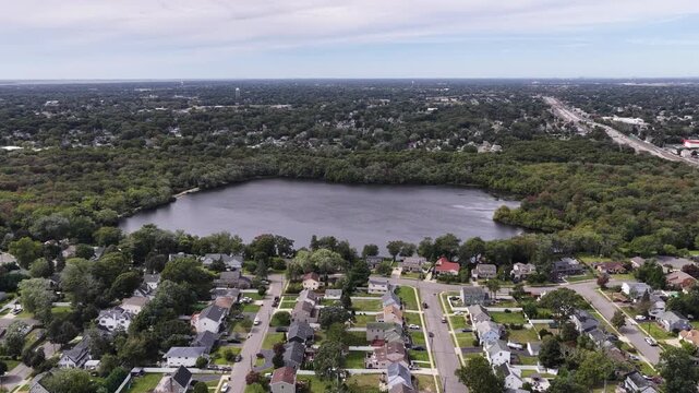 A drone approaches Southards Pond in Babylon on Long Island in New York. The freshwater pond sits in dense woodland beside residential streets and detached homes with grassy lawns and leafy yards.