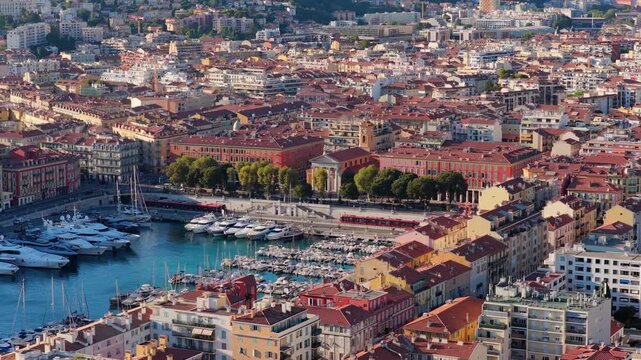 Slow tele orbit over Port Lympia marina with luxury yachts, red trams and Notre Dame du Port church in warm afternoon sun, Nice, French Riviera, Cote d Azur, France