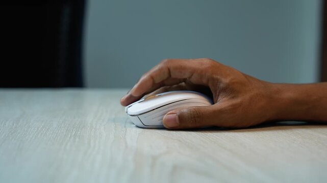 Hand Using Wireless Mouse on Wooden Desk, Close-up of a hand using a white wireless computer mouse on a light wooden desk, with a blurred dark and blue background.