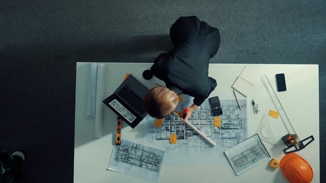 Top view of skilled project manager holding project plan and explain house design to architect engineer at meeting table with equipment, ruler, safety helmet and laptop scatter around. Alimentation.