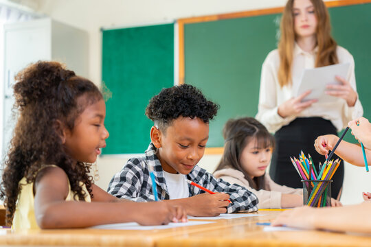 Multiethnic school children drawing with colored pencils in classroom with teacher, diverse students learning art and writing at desk with educator supervision
