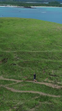 Solo woman explores scenic coast.Woman treks solitary over vibrant seaside ridge during summer.Solitary female hiker traverses winding grassy route overlooking tranquil turquoise waters,vertical video