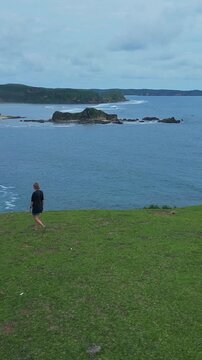 Woman gazing seaward. Solo woman pauses by ocean edge. Solitary woman stops at grassy cliff overlook. Individual female traveler gazes across calm ocean waters from rocky precipice, vertical video