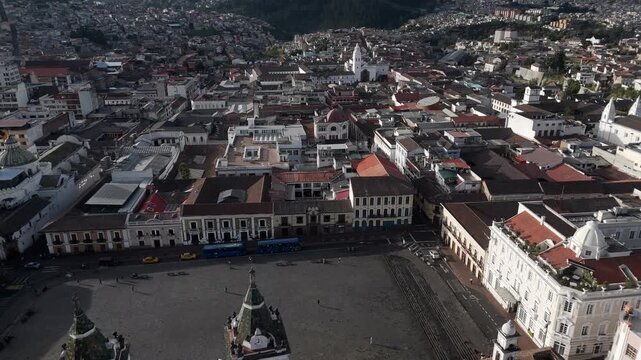 Quito's historic center unfolds from above. White towers flank a central plaza. Red-tiled roofs stretch across the town. Sunlight bathes ancient stone buildings. People move calmly through the streets