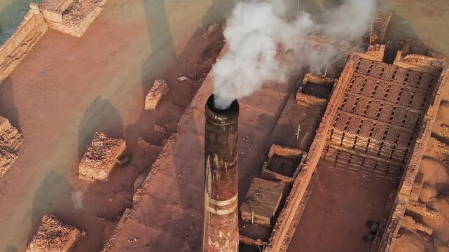 Aerial view of a brick kiln in Bangladesh with a tall chimney releasing dense smoke above stacks of fired clay bricks, highlighting traditional production and industrial air pollution.