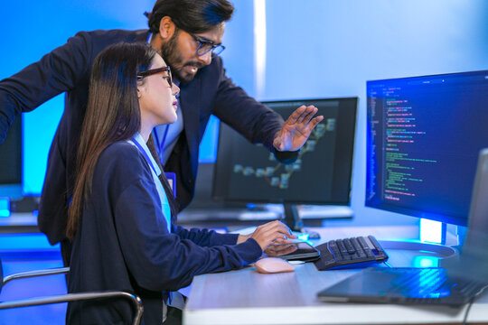A male engineer and a female colleague discussing source code shown on the monitor, working together at a coding workstation in a blue-lit office; candid moment of software development.