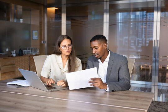 Two multiethnic business colleagues working at office desk, preparing report, reviewing project plan, discussing document terms, working on agreement details during corporate teamwork in meeting room