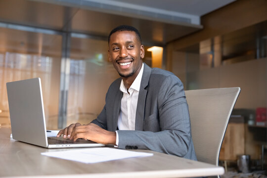 Portrait of smiling African businessman working on project, preparing report, typing on laptop sitting at workplace desk in corporate office. Professional business workflow, technology, executive work