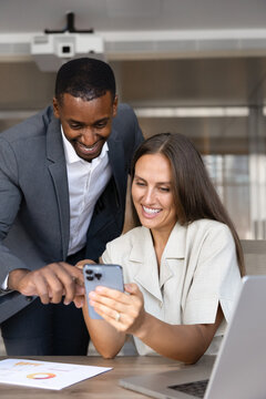 Smiling man showing to woman colleague new professional mobile app on smartphone at teamwork meeting in corporate office. Digital workflow, platform discussion, positive business information review