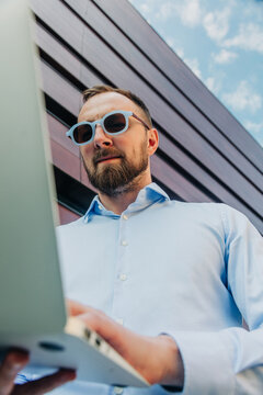 Businessman in stylish sunglasses working on laptop outside office, remote work