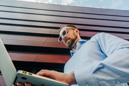 Businessman in stylish sunglasses working on laptop outside office, remote work