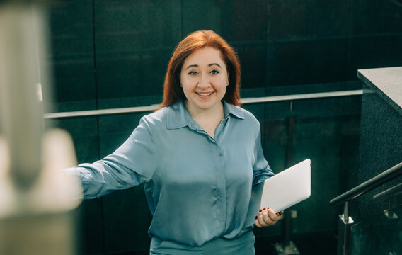 Redhead businesswoman in blue suit with laptop walking up office stairs