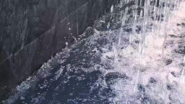 Cascading water curtain falling into stone pool wall creating sparkling ripples and foam bubbles
