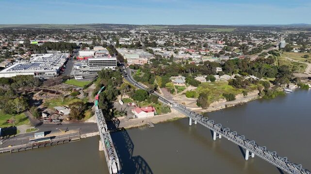 Aerial drone view of the historic steel truss road and rail bridges crossing the Murray River at Murray Bridge, South Australia, with the regional town centre and commercial buildings beyond.