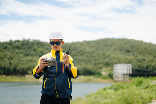 Professional engineer wearing a hard hat operates a drone remote while talking on a walkie-talkie. Outdoor site inspection near a scenic lake