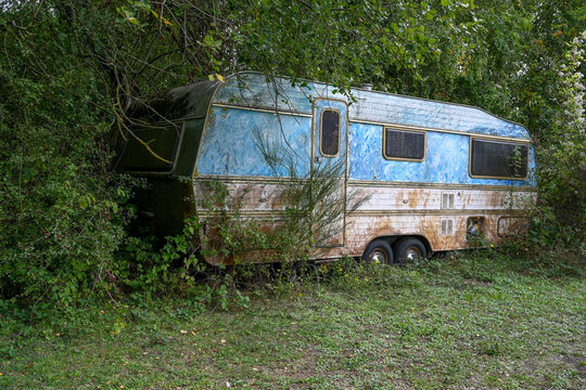 une vieille caravane rouill&eacute;e est abandonn&eacute;e et recouverte par la v&eacute;g&eacute;tation sauvage. Une fa&ccedil;ade de l'abri de camping est bleue et blanche, avec la porte, les fen&ecirc;tres et les roues visibles