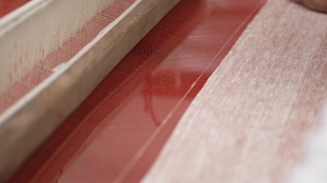 Close up of artisan's hands working the beater on a traditional wooden loom to weave red silk fabric.