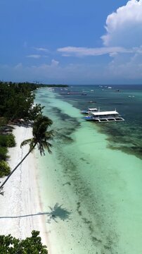 Vertical drone ascends while flying over Doljo Beach, showing white sand, turquoise shallows, outrigger boats, palms, and the Panglao coastline