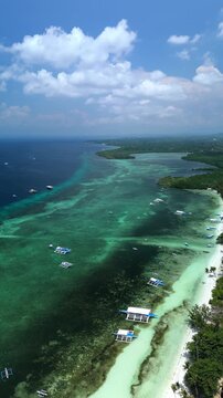 Vertical drone pans left during a flyover, revealing Doljo Beach, turquoise reef waters, boats, white sand, and coastline in Panglao, Bohol