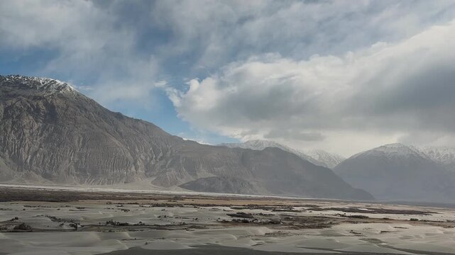 tilt up view: grey sand dunes surrounded by mountain range under cloudy blue sky in himalayas