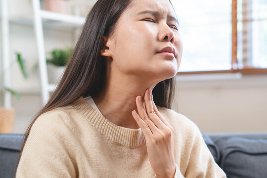 Young Asian woman suffering from sore throat and touching her neck with hand at home, feeling sick with a cup of hot tea or warm water, healthcare and medical problem concept