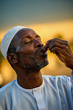 Black man in a white kufi about to eat a date at sunset