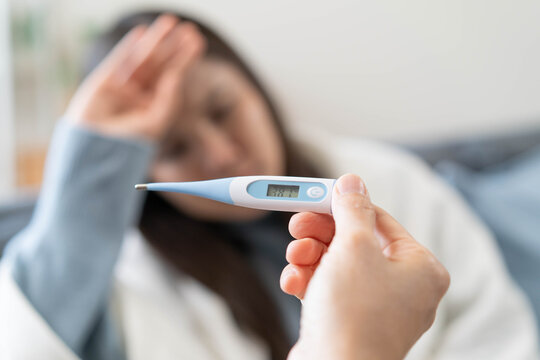 Sick person resting in bed checking temperature with thermometer, suffering from high fever, seasonal flu, influenza virus infection, and common cold symptoms at home