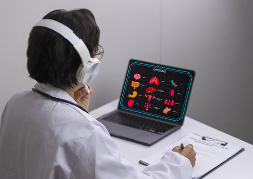 Doctor headphone white coat and stethoscope studying human organ diagram on laptop taking notes on clipboard medical education modern technology focused learning