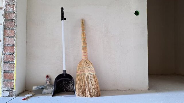 Broom and dustpan standing in an unfinished room during renovation, with exposed brickwork, plastered walls, construction materials, and a bare concrete floor