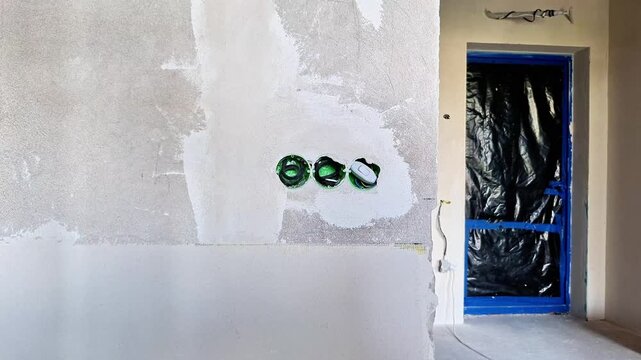 Unfinished interior wall with three electrical outlet boxes installed during renovation, featuring rough plaster, exposed wiring, and a protected doorway under construction