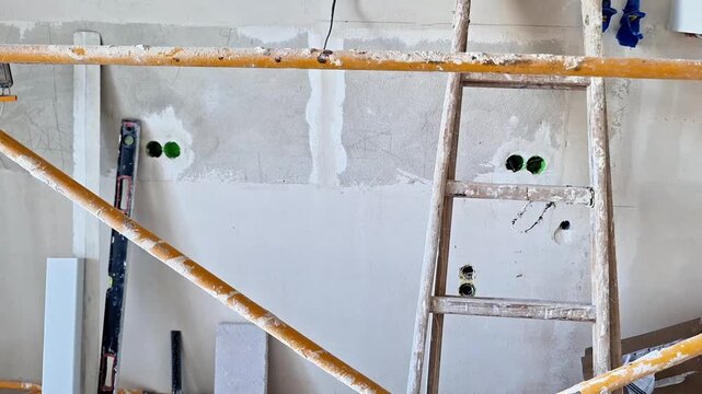 Unfinished interior wall during renovation with electrical outlet boxes, exposed wiring, plasterwork, wooden supports, and construction tools visible on site